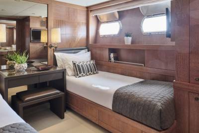 Two neatly made twin beds with patterned pillows and quilted blankets fill the foreground, while the background features wood-paneled walls, porthole windows, and a television.