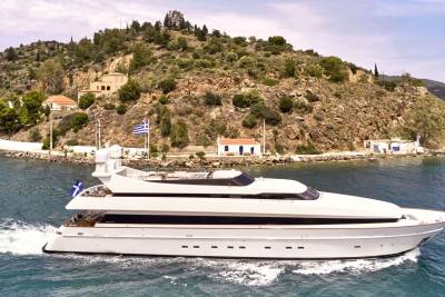 A sleek white yacht cruises through blue water in the foreground, with a picturesque Greek island dotted with white buildings and lush greenery in the background.