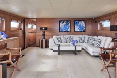 Two wooden chairs with striped side tables are in the foreground, while a large white sectional sofa with blue and white patterned pillows sits in the background, facing two framed blue floral artworks on a wood-paneled wall.
