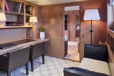 A wooden desk with two chairs sits in the foreground, while a hallway with patterned tiles leads to a bedroom in the background.