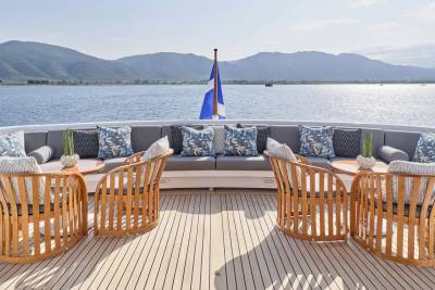 Wooden chairs and plush seating arrangements on a yacht's deck face a calm blue sea and distant, hazy mountains under a bright sky.