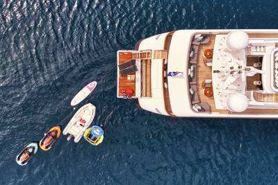 A large yacht floats on the deep blue sea, with several inflatable water toys trailing behind it.