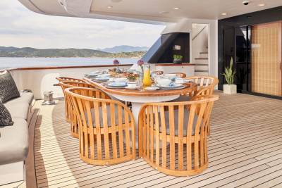 A beautifully set dining table with chairs on a yacht's deck overlooks a serene coastal landscape.