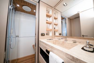A modern bathroom with a marble vanity and sink in the foreground and a glass-enclosed shower with a wooden slatted floor in the background.