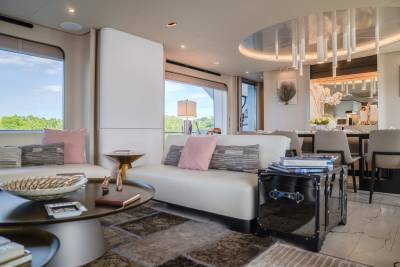 A luxurious yacht interior features a white sofa and decorative trunk in the foreground, with a dining area and large windows overlooking greenery in the background.