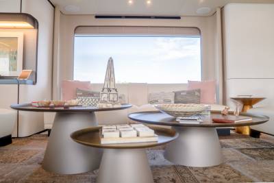 Three modern, metallic coffee tables with decorative items sit in the foreground of a luxurious living area with a large window offering a view of the sky in the background.