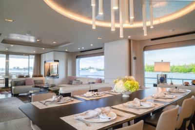 A long dining table set for a meal occupies the foreground, while a spacious living area with sofas and large windows overlooking a body of water with lush greenery forms the background.