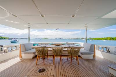 A dining area with a table and chairs on a yacht's deck overlooks a calm bay dotted with boats under a bright sky.