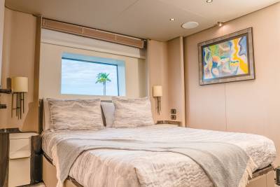 A neatly made bed with plush pillows and a light gray throw occupies the foreground, while a framed abstract painting hangs on the wall above a nightstand in the background, with a window revealing a palm tree and sky.