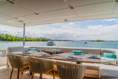 A long dining table with place settings is set on a yacht's deck, overlooking a calm bay dotted with boats and a lush green coastline under a bright blue sky.