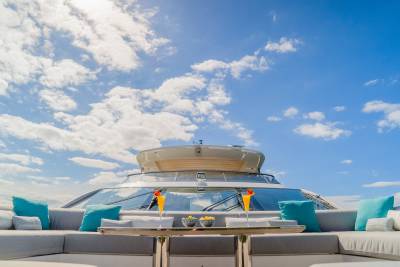Two cocktails and snacks rest on a table on a yacht's deck, with a bright blue sky and scattered clouds in the background.
