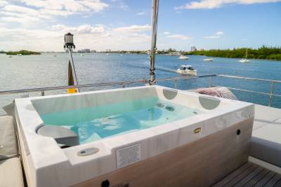 A luxurious hot tub filled with clear blue water sits on a yacht's deck, with a calm bay dotted with boats and lush green islands in the background.