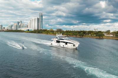 A sleek white yacht cruises on a wide river, leaving a foamy wake, with a city skyline and lush trees lining the distant shore under a dramatic cloudy sky.