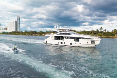A sleek white yacht cruises on a calm waterway, leaving a foamy wake behind a jet ski, with a city skyline and palm trees visible in the distance under a cloudy sky.