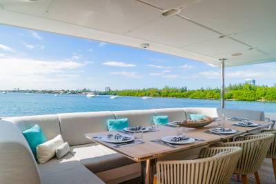 A dining table set for a meal on a yacht's deck overlooks a calm blue bay dotted with boats and lined with lush green trees under a bright sky.