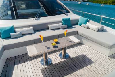 Two cocktails and a bowl of fruit sit on a table in the foreground of a luxurious yacht's seating area, with a calm blue ocean and distant boats in the background.