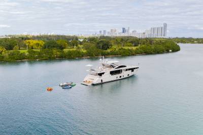 A large white yacht with smaller dinghies attached floats on calm blue water, with a lush green coastline and a distant city skyline under a cloudy sky.