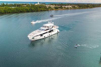 A large white yacht cruises on a wide blue waterway in the foreground, with a jet ski and two smaller boats trailing behind, while a lush green coastline with buildings and a lighthouse forms the background.