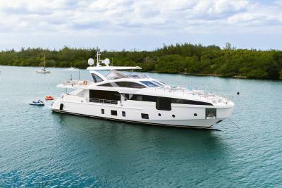 A sleek white yacht floats on calm blue water in the foreground, with lush green trees and a cloudy sky forming the background.