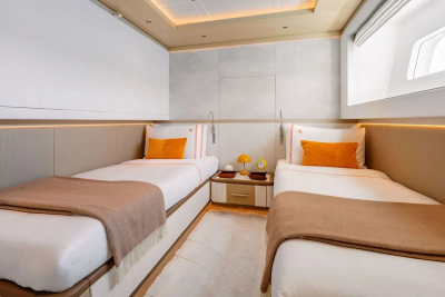 Two neatly made twin beds with orange pillows and brown throws occupy the foreground, while a nightstand with a lamp and a window are visible in the background.
