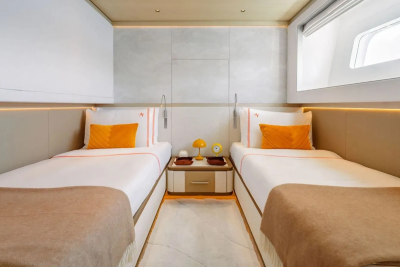 Two neatly made twin beds with orange pillows and brown throws are in the foreground, with a nightstand between them, while the background features a textured wall and a window with a curved frame.
