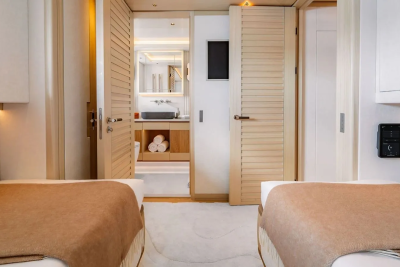 Two beds with brown blankets are in the foreground, leading to a bathroom with a sink and towels in the background.