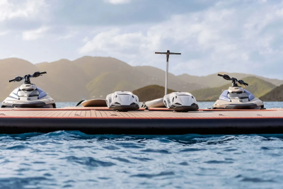 Four jet skis are parked on a floating dock in the foreground, with lush green hills and a cloudy sky in the background.