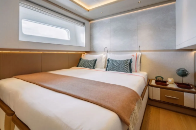 A neatly made bed with patterned pillows and a rust-colored throw dominates the foreground, while a modern nightstand with decorative items sits to the right, all set against a backdrop of textured walls and a large window with a view of the sky.