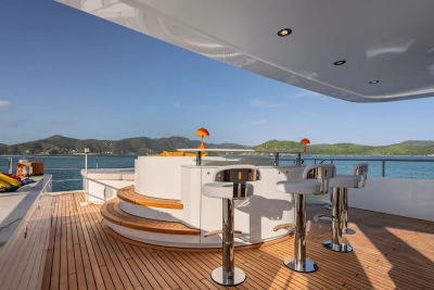 A luxurious yacht deck with a bar and stools in the foreground overlooks a serene ocean and distant, verdant islands under a clear blue sky.