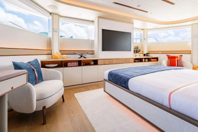A luxurious bedroom with a plush armchair and a large bed in the foreground, and a wall-mounted television and expansive windows overlooking a bright sky in the background.