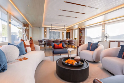 Plush white sofas and armchairs with blue and orange accents fill the foreground of a luxurious yacht's living area, while a long dining table and chairs are visible in the background, all illuminated by soft, recessed lighting and natural light from large windows.