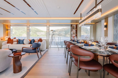 A luxurious yacht interior features a dining table set for a meal in the foreground and a comfortable seating area with large windows overlooking a scenic landscape in the background.