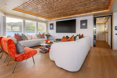 In the foreground, two vibrant orange chairs face a spacious white sofa, while the background reveals a large television and a window overlooking a serene body of water.