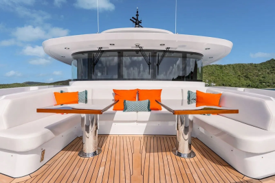 A luxurious yacht's deck with white seating and orange cushions in the foreground, set against a backdrop of lush green hills under a clear blue sky.