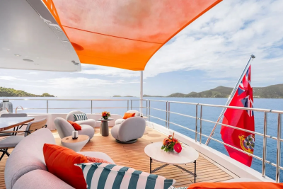A luxurious yacht deck with comfortable seating and vibrant cushions in the foreground is set against a backdrop of a calm blue sea and lush green islands under a bright, cloudy sky.