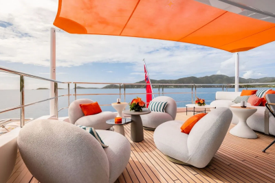 Plush white lounge chairs with orange accents are arranged on a wooden deck under an orange canopy, with a calm blue sea and distant green hills in the background.