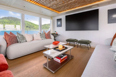 A modern living room with plush sofas and coffee tables in the foreground overlooks a serene ocean and lush green hills in the background.