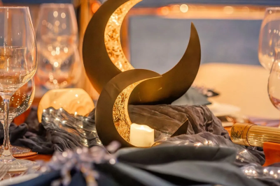 A candle glows within a crescent moon decoration on a table set with wine glasses and fairy lights, with a blurred background of more glassware and warm lighting.