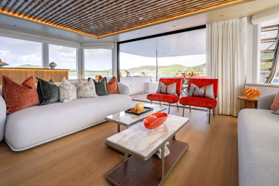A luxurious yacht interior with plush white sofas and colorful accent chairs in the foreground, and a scenic view of rolling hills and water through large windows in the background.