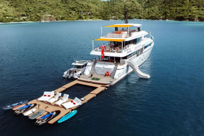 A large white yacht with a slide is docked at a floating platform with lounge chairs and water toys, set against a backdrop of lush green hills and calm blue water.