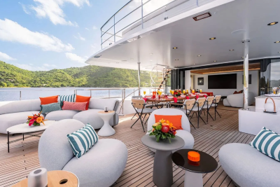 Plush white seating arrangements and a dining table are set on a yacht's deck, with a lush, green, hilly coastline visible in the background under a bright blue sky.