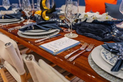 A formal dining table is set with elegant place settings and floral arrangements on a yacht, with blue cushions and pillows visible in the background.