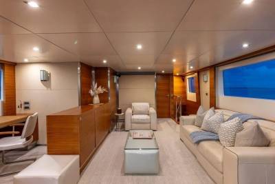A modern yacht interior features a cream sofa and armchairs in the foreground, with large windows revealing a blue ocean and sky in the background.