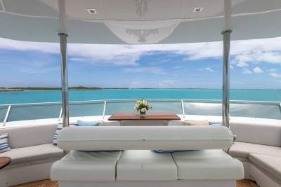 A white cushioned seating area with a wooden table holding a floral arrangement is in the foreground, overlooking a calm turquoise ocean under a bright blue sky in the background.