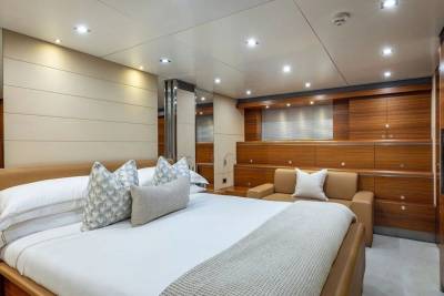 A luxurious yacht bedroom features a large bed with decorative pillows in the foreground and a seating area with wooden accents in the background.