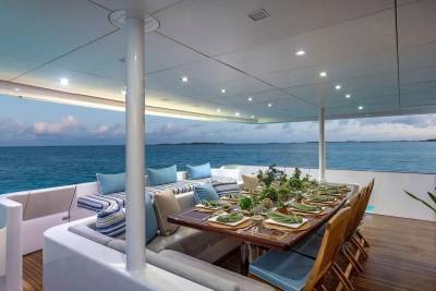 A beautifully set dining table on a yacht's deck is surrounded by plush seating, with the calm blue ocean and distant islands visible in the background.