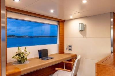 A laptop and flower arrangement sit on a desk in front of a large window overlooking a calm sea and distant islands.