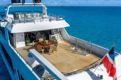 A luxurious yacht with an outdoor dining area in the foreground sails on a calm, turquoise ocean under a clear sky.