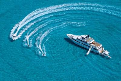 A large white yacht floats in clear turquoise water, while several jet skis create swirling wakes around it.