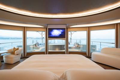 A luxurious bedroom with a large white bed in the foreground overlooks a serene ocean view through large windows, with a television screen mounted above a hot tub in the background.